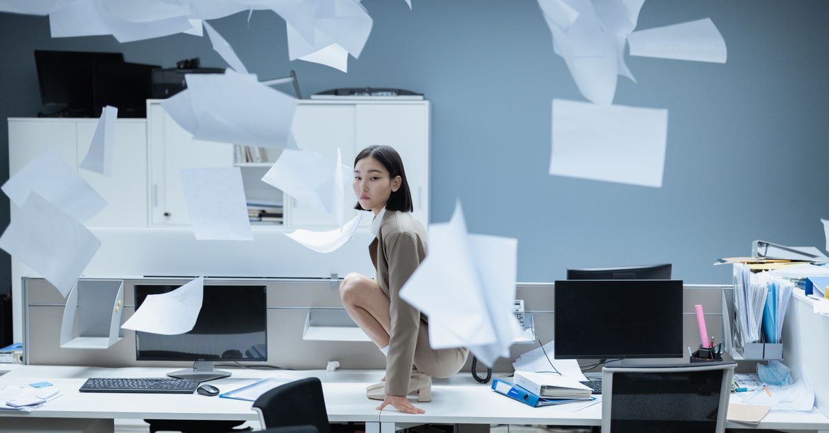Stop Doing Repetitive Work: AI Workflow Automation Strategies for London Businesses Woman perched on a desk in a chaotic office with papers flying, representing workplace stress.